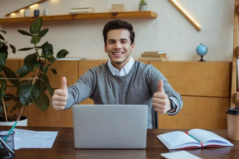 Businessman with thumbs up smiling and looking at camera near laptop at table in office
